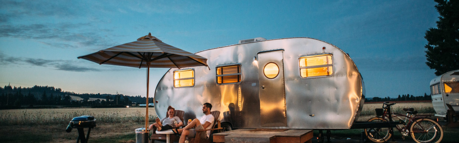 A couple relaxes in chairs under an umbrella outside a shiny silver camper at dusk, with warm lights glowing inside. A couple relaxes in chairs under an umbrella outside a shiny silver camper at dusk, with warm lights glowing inside.