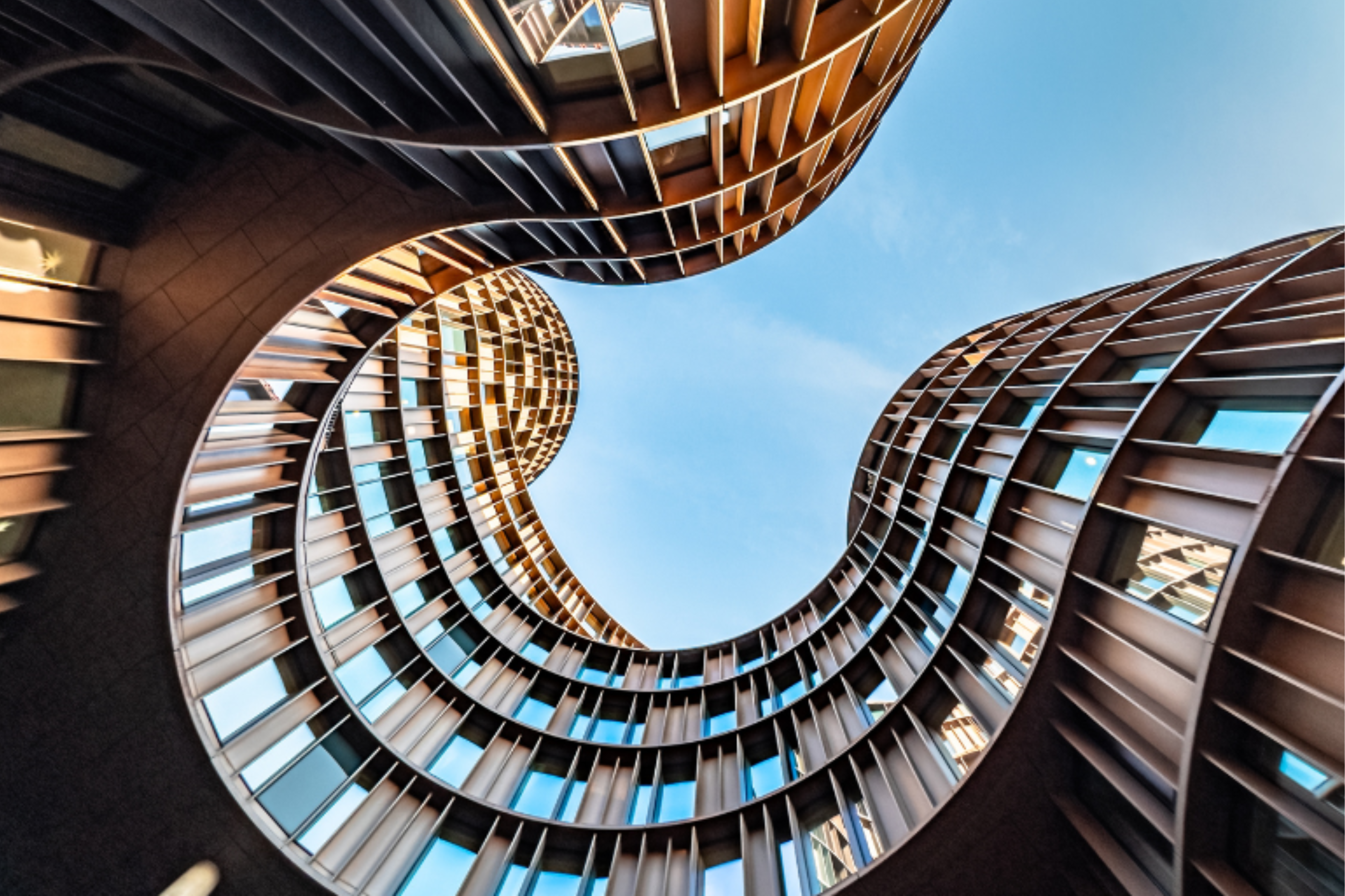 Upward view of a modern building with curved, wave-like architectural forms and reflective windows framing the blue sky.
