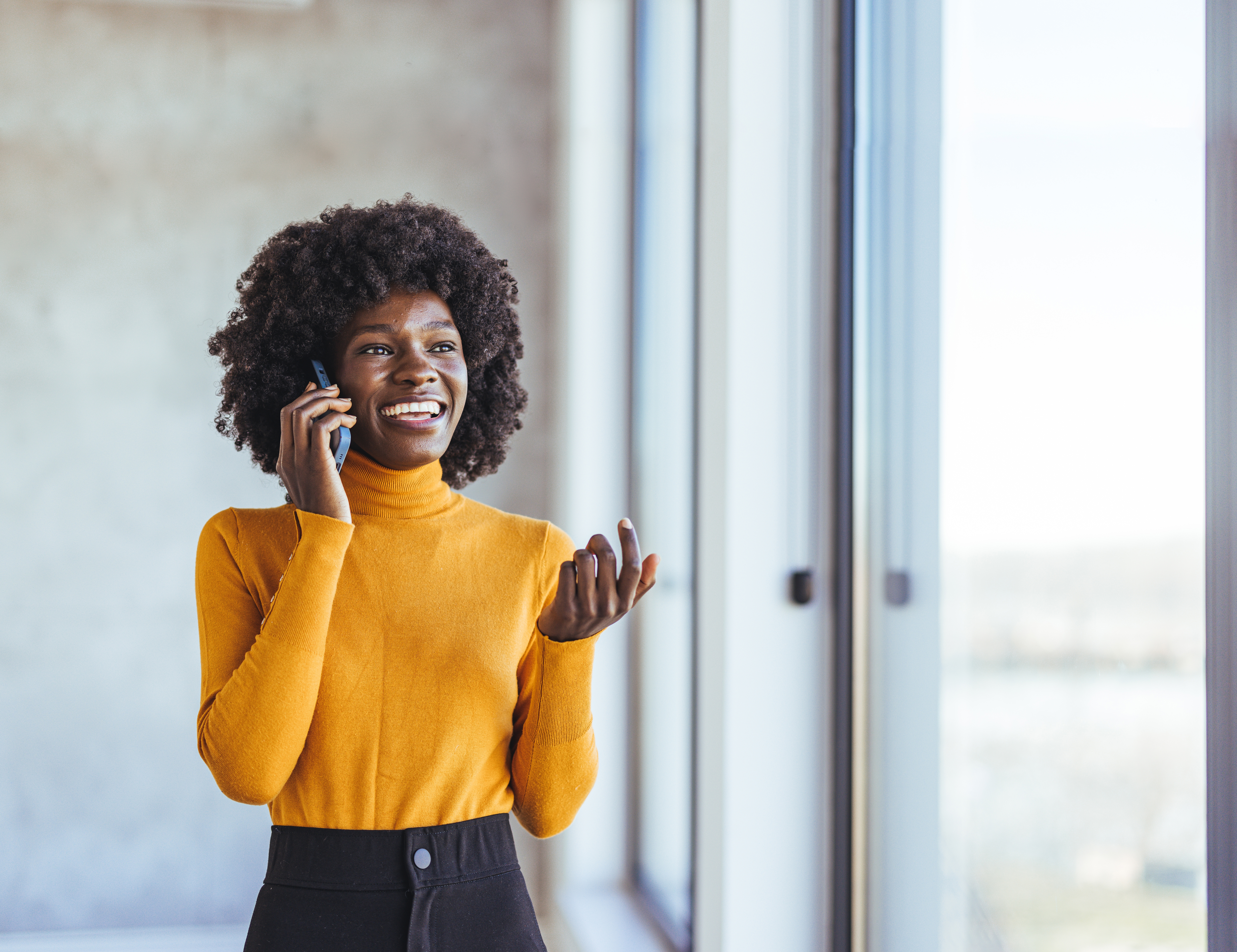 Smiling woman talking on phone near window, wearing yellow sweater