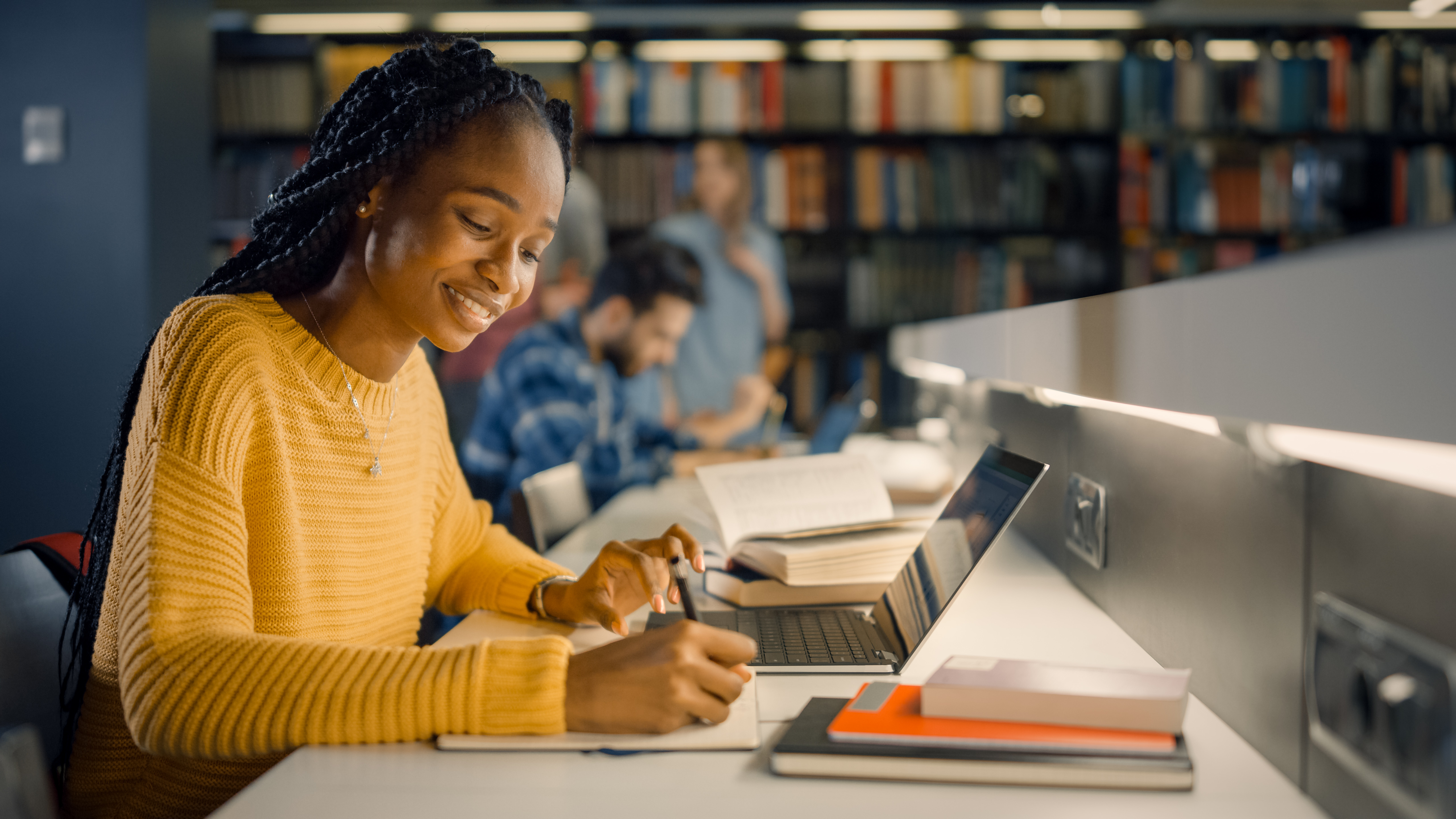 Student studying at library desk using laptop and taking notes