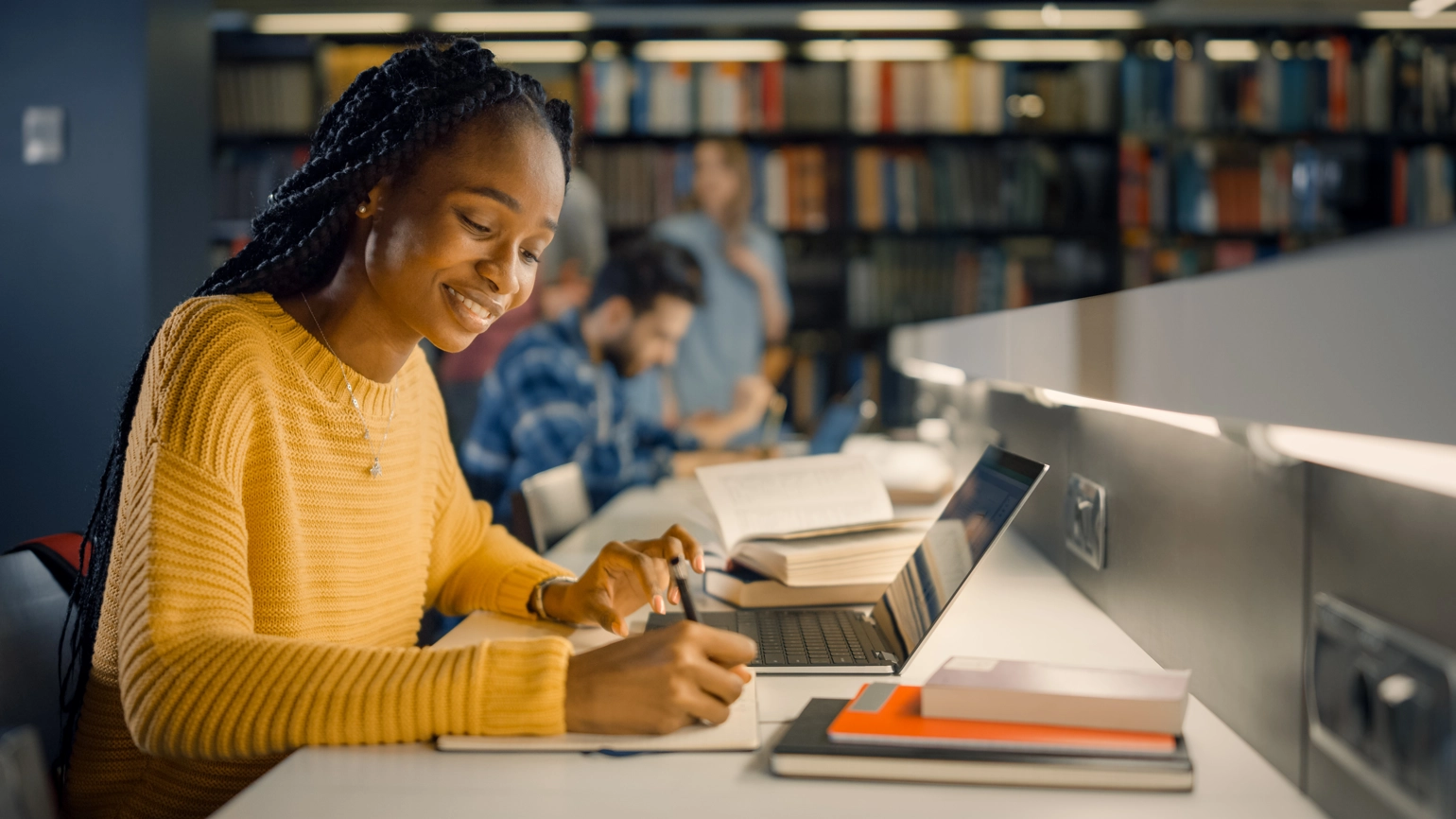 Student studying at library desk using laptop and taking notes Student studying at library desk using laptop and taking notes