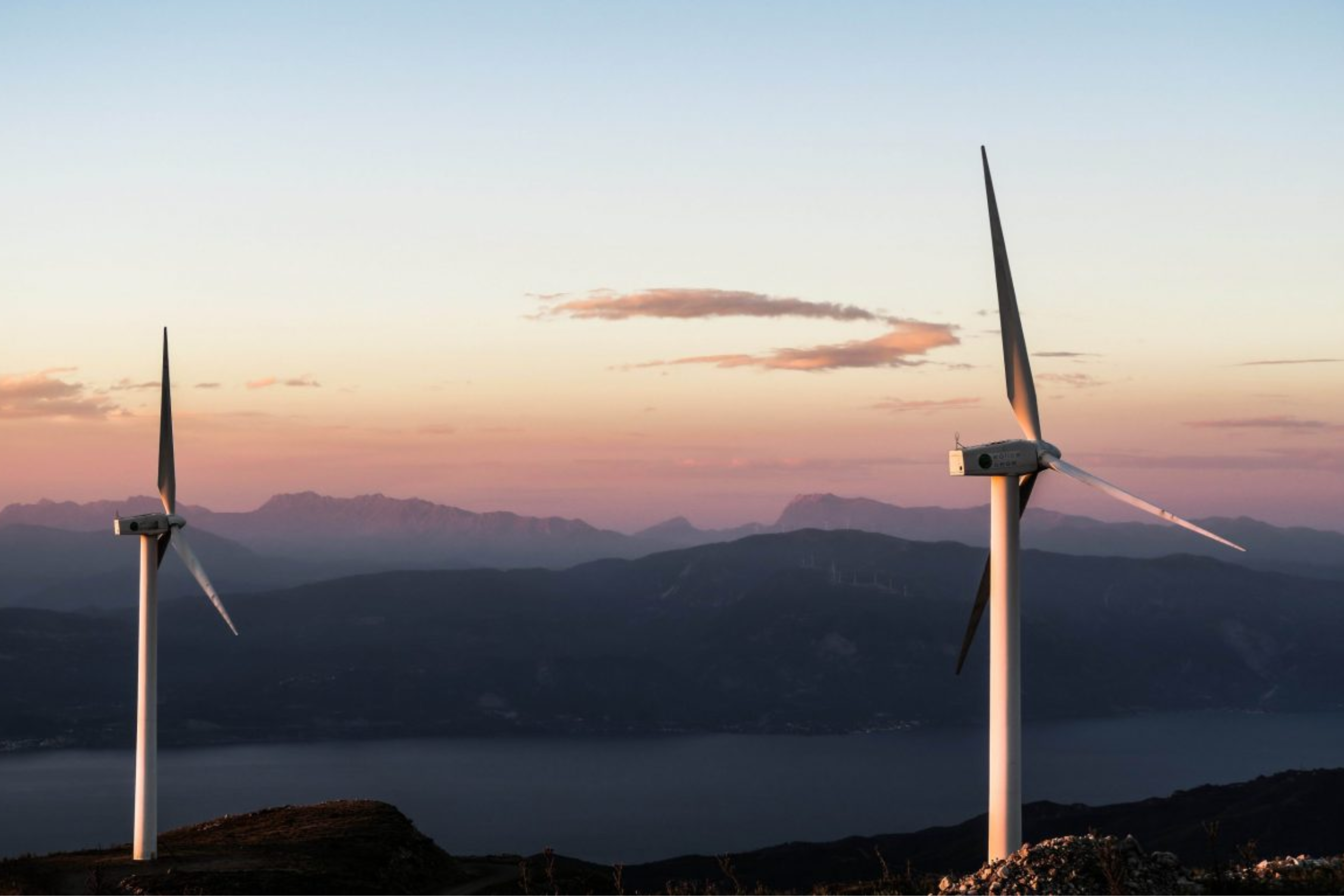 Two wind turbines standing on a hillside at sunset with mountains in the background.