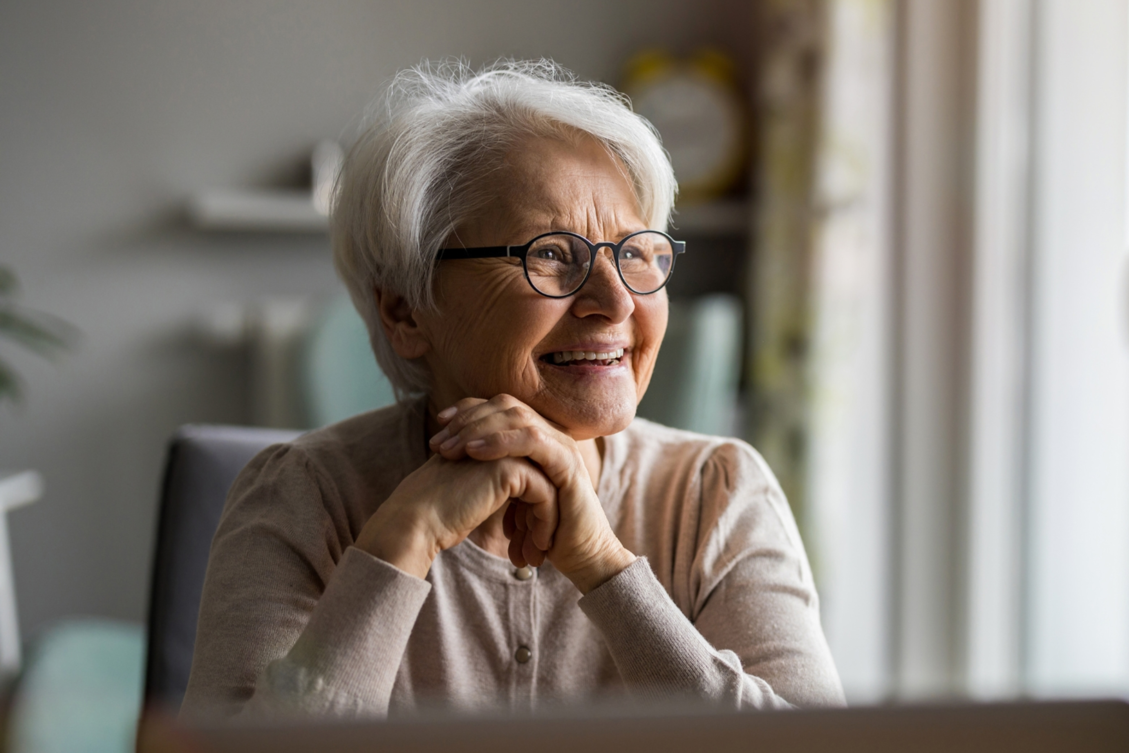 Smiling older woman with short white hair and glasses looking out a window with her hands clasped under her chin.