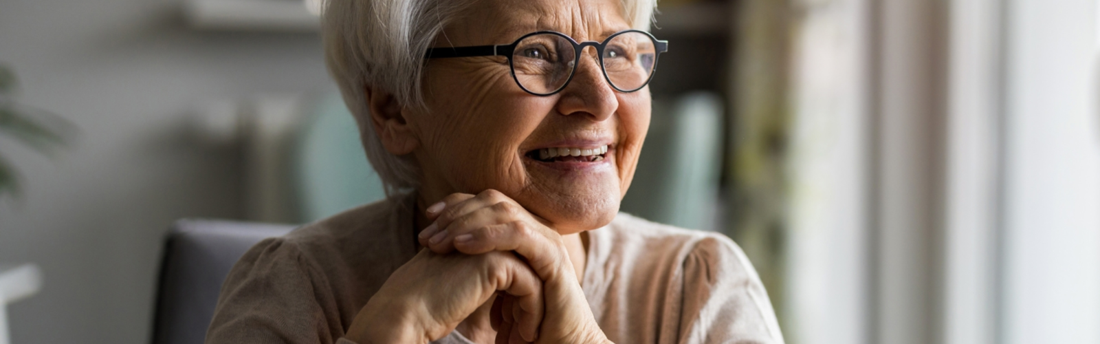 Smiling older woman with short white hair and glasses looking out a window with her hands clasped under her chin. Smiling older woman with short white hair and glasses looking out a window with her hands clasped under her chin.