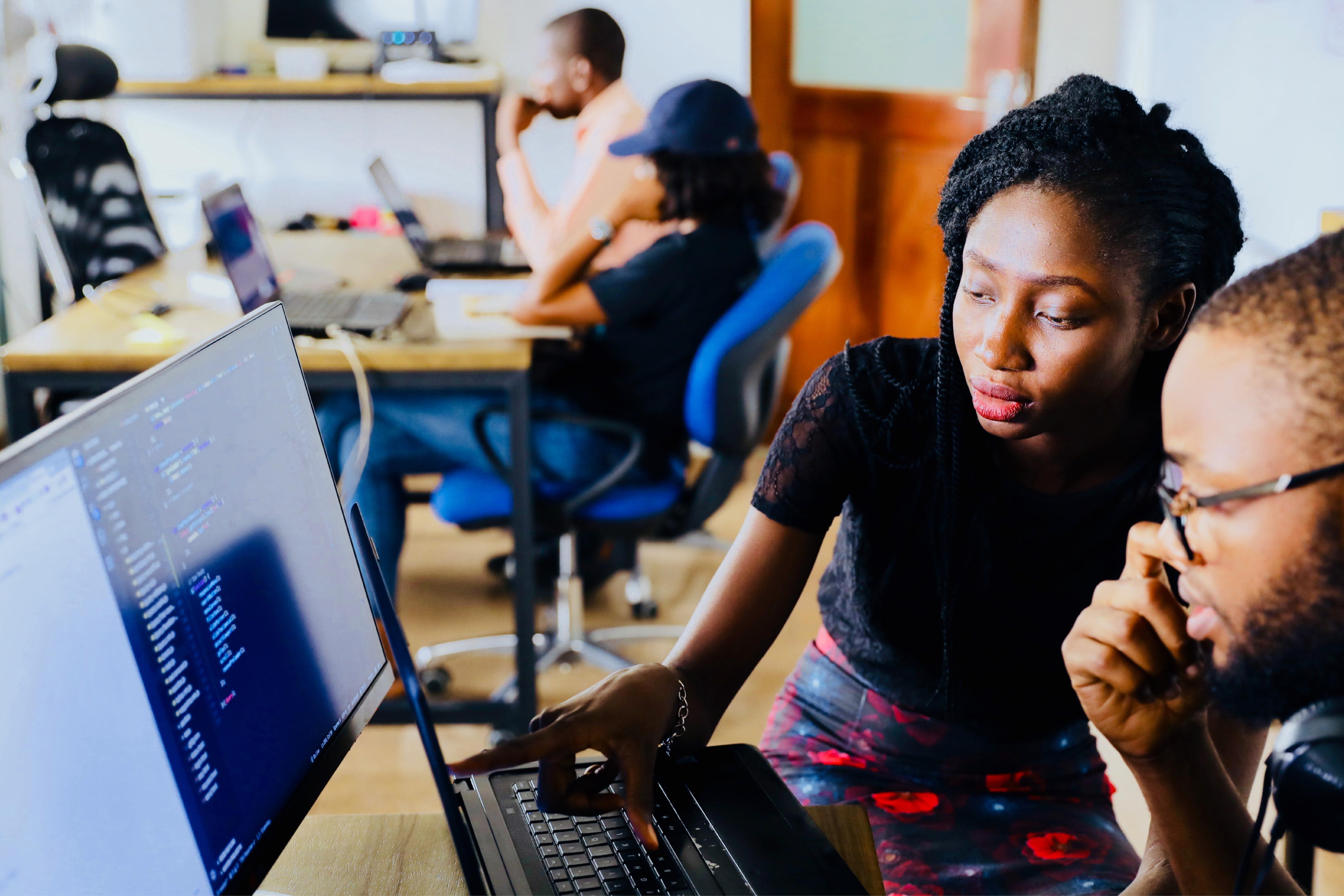 A woman points at code on a laptop screen while working with a colleague in a shared office space