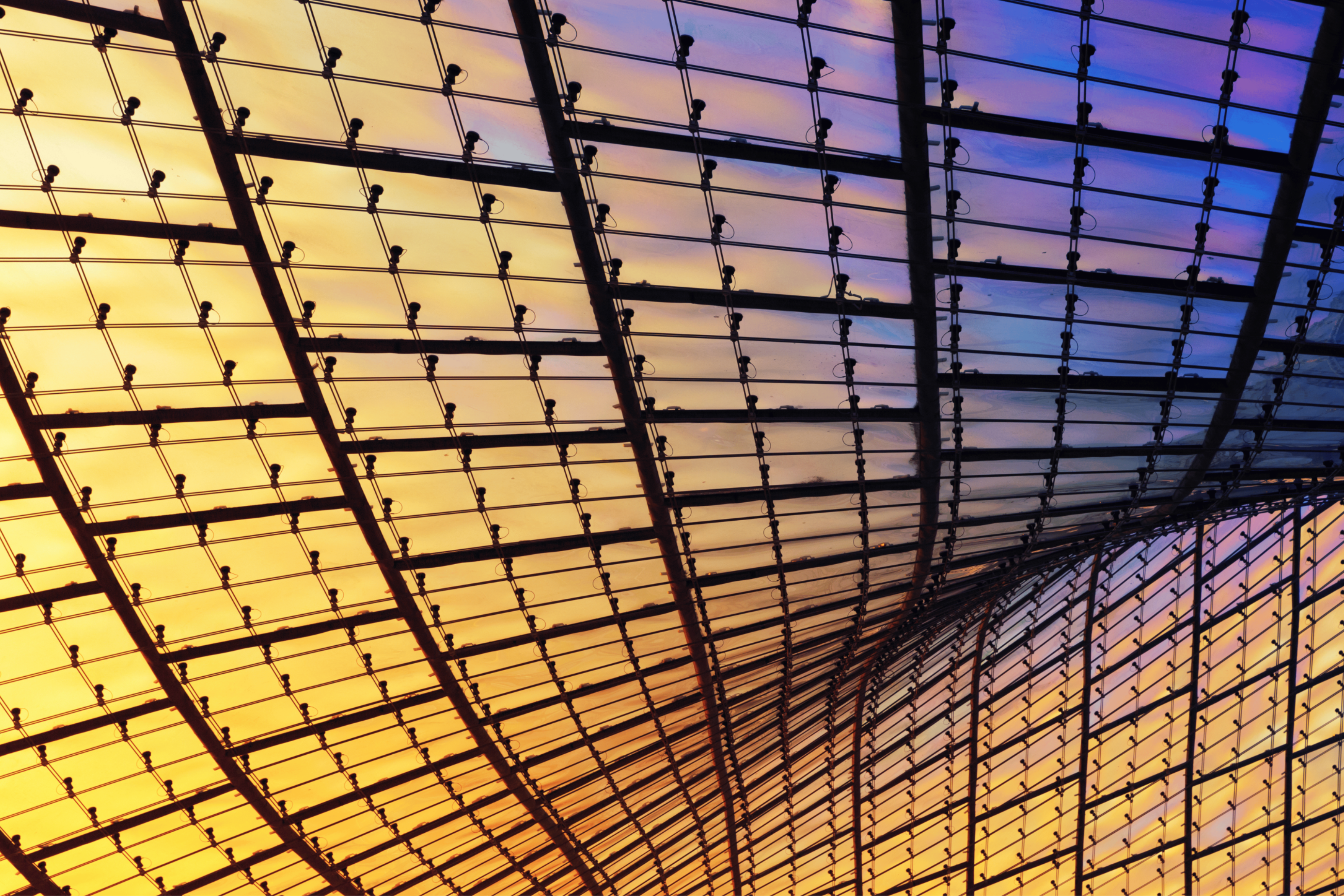 Curved glass and steel ceiling structure glowing with warm yellow and cool purple light.