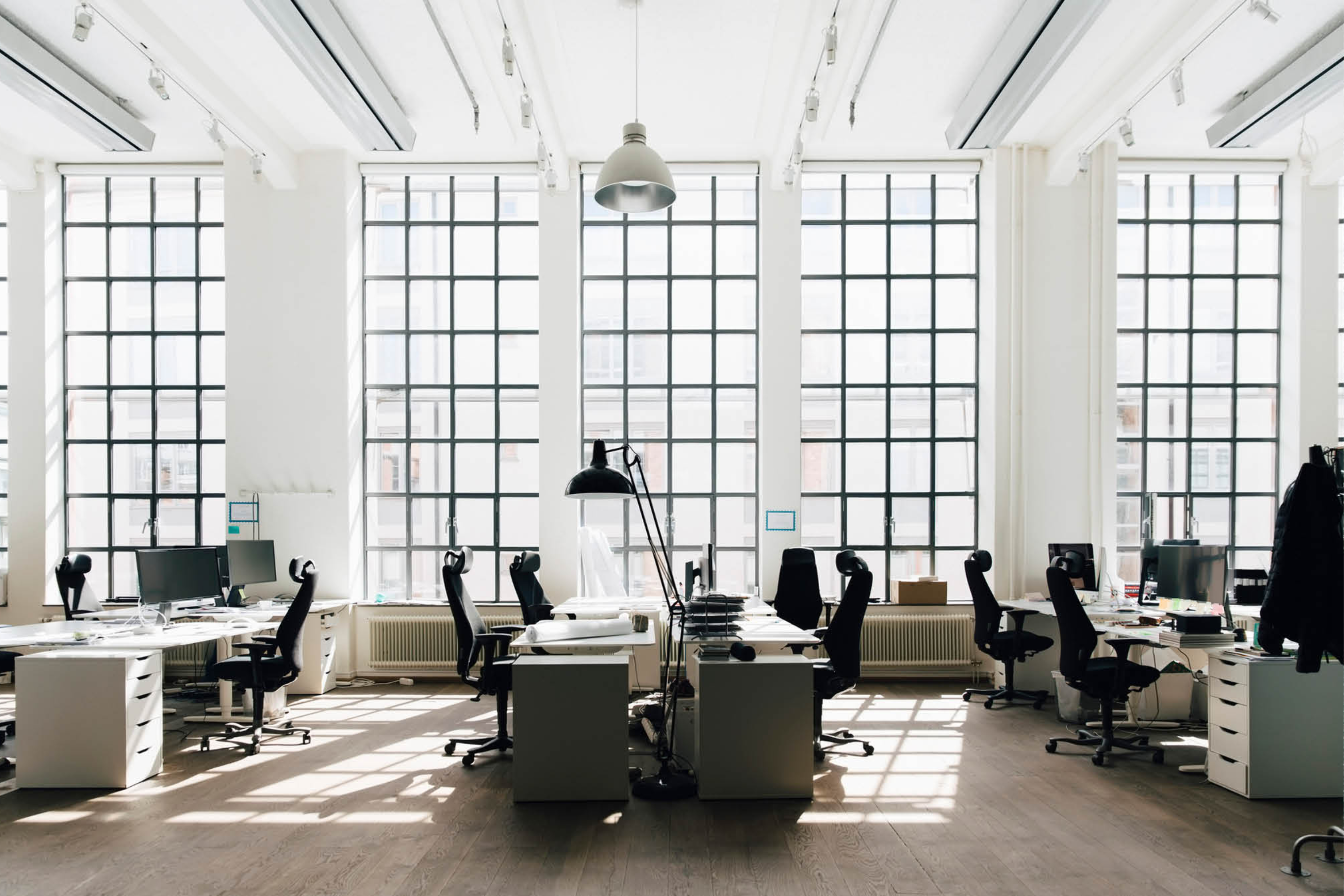 A bright, modern office space with large grid windows, empty desks, and black office chairs arranged in rows.