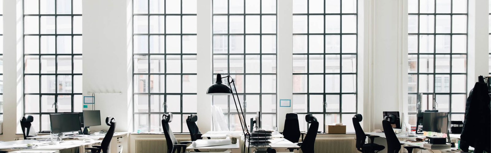 A bright, modern office space with large grid windows, empty desks, and black office chairs arranged in rows. A bright, modern office space with large grid windows, empty desks, and black office chairs arranged in rows.