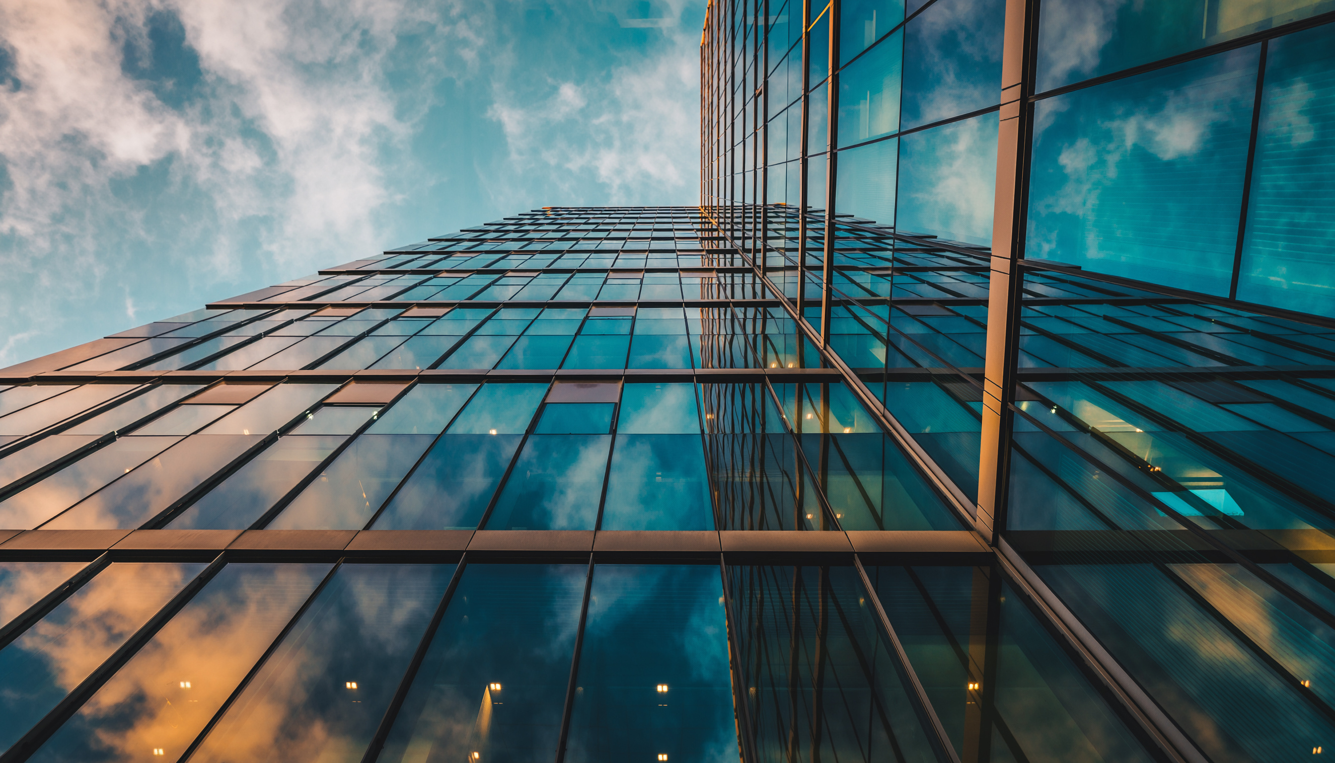 Upward view of modern glass skyscraper reflecting blue sky and clouds, with geometric lines and warm interior lights at dusk.