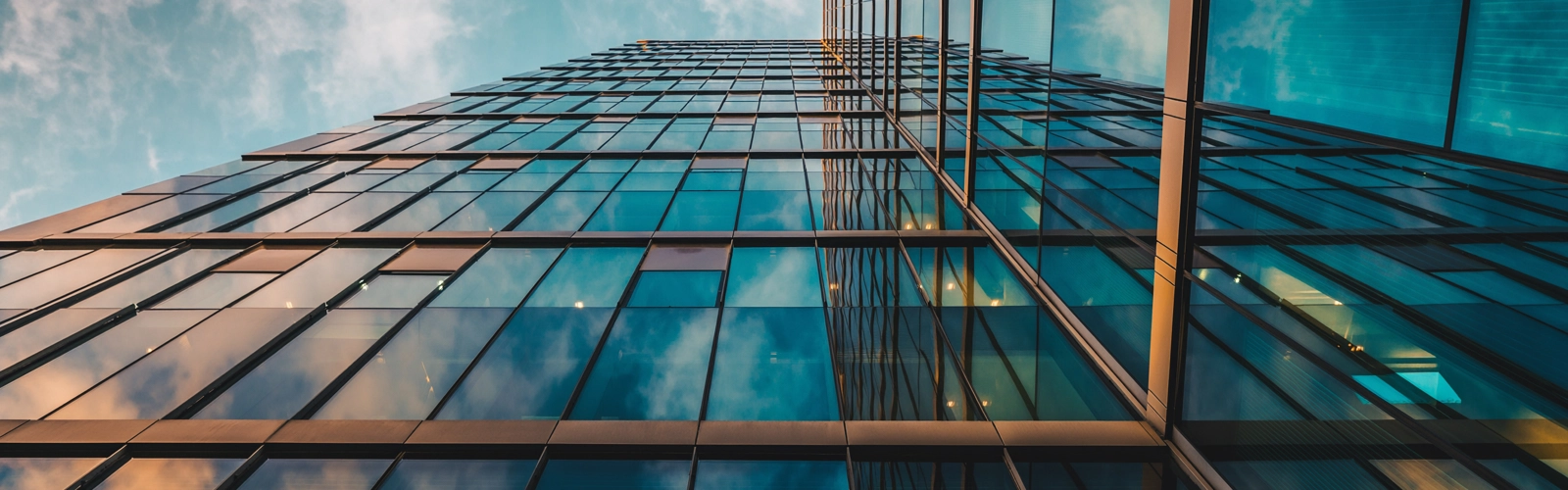 Upward view of modern glass skyscraper reflecting blue sky and clouds, with geometric lines and warm interior lights at dusk. Upward view of modern glass skyscraper reflecting blue sky and clouds, with geometric lines and warm interior lights at dusk.