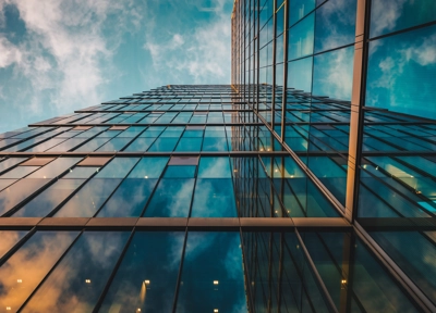 Upward view of modern glass skyscraper reflecting blue sky and clouds, with geometric lines and warm interior lights at dusk. Upward view of modern glass skyscraper reflecting blue sky and clouds, with geometric lines and warm interior lights at dusk.