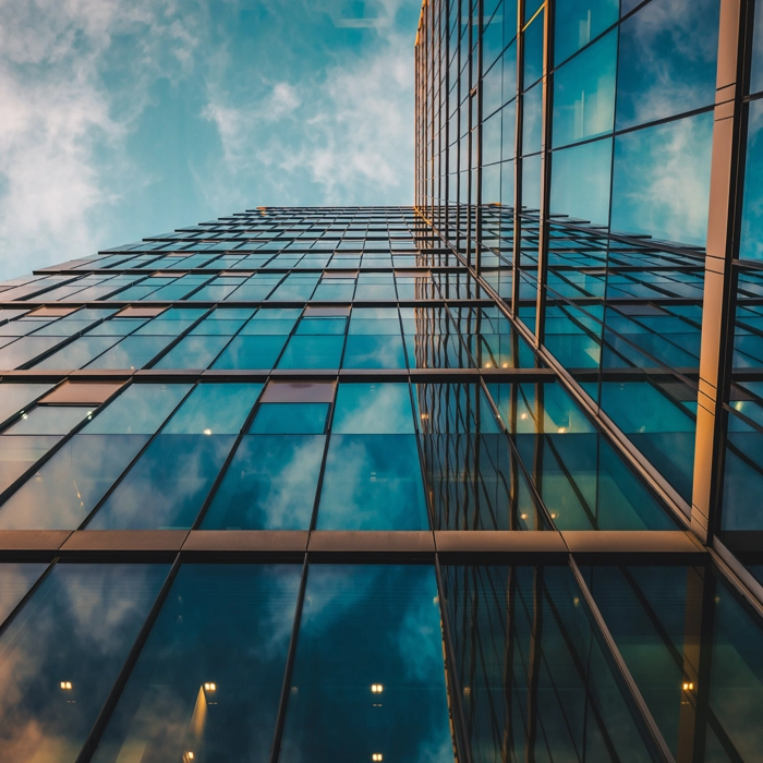 Upward view of modern glass skyscraper reflecting blue sky and clouds, with geometric lines and warm interior lights at dusk. Upward view of modern glass skyscraper reflecting blue sky and clouds, with geometric lines and warm interior lights at dusk.